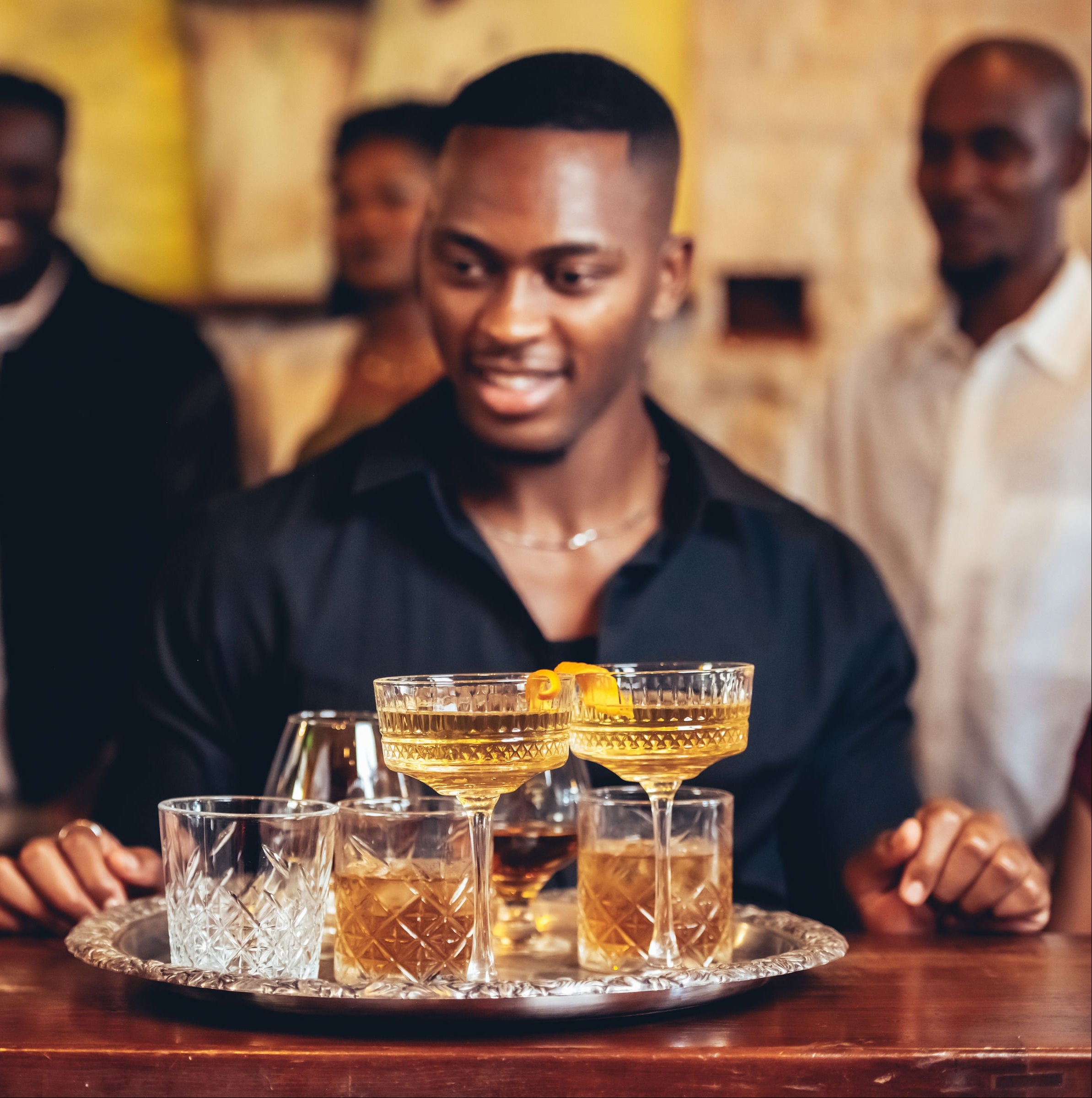 Man holding a bottle of Avante X.O. brandy with a tray of cocktails in front of him, surrounded by friends.